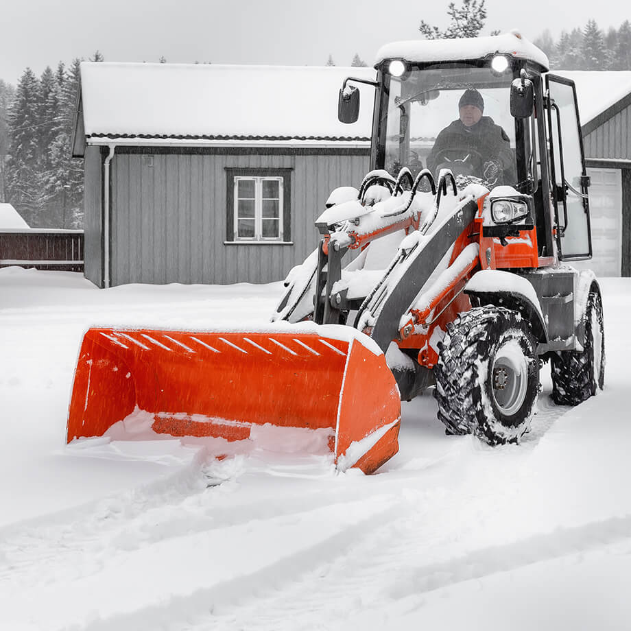 A person operates an orange snow plow during a heavy snowfall, clearing a path on a snow-covered landscape with a grey wooden house in the background.
