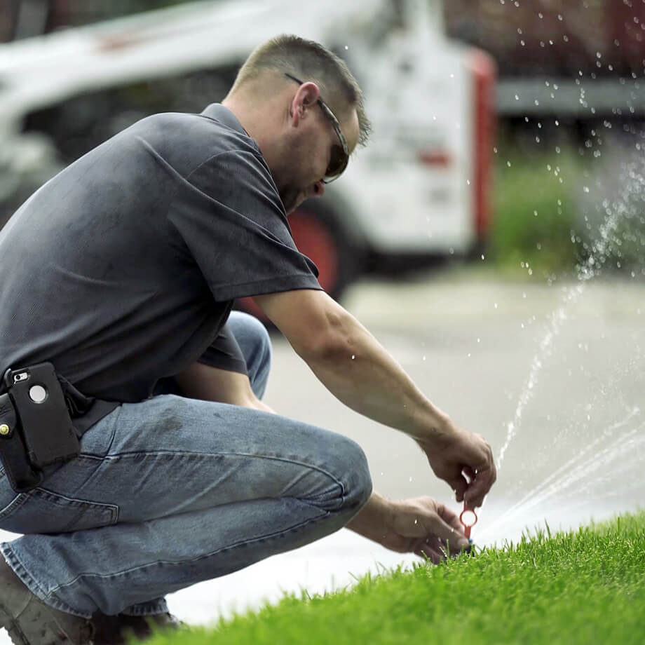 A technician in casual work attire is crouched down adjusting a sprinkler head with water spraying out onto a lush green lawn, with a utility van in the background.
