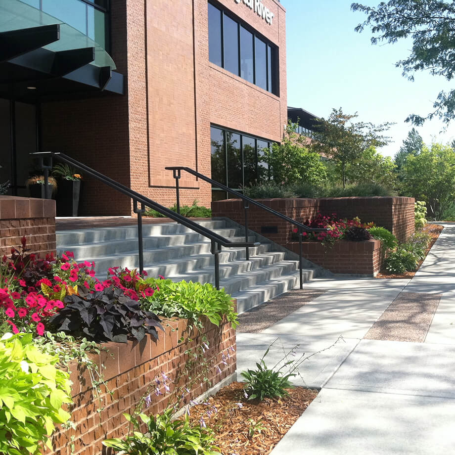 Entrance of a modern brick office building with large windows, featuring landscaped beds with vibrant pink flowers and lush green plants along the sidewalk.